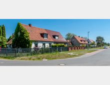 Straßenszene in einer Wohnsiedlung mit Einfamilienhäusern, die rote Ziegeldächer und teilweise bewachsene Fassaden haben. Die Häuser sind von Gärten mit Zäunen umgeben, und eine asphaltierte Straße mit Gehweg führt daran vorbei.