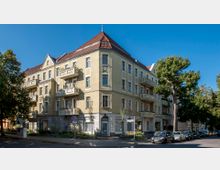 Eckgebäude im Stil eines Altbaus mit drei Stockwerken, gelber Putzfassade und roten Ziegeldächern, großen Fenstern sowie mehreren Balkonen. Im Vordergrund sind Bäume, geparkte Autos und eine Straßenkreuzung zu sehen.