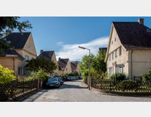 Gepflasterte Wohnstraße mit symmetrischen Reihenhäusern, die cremefarbene Fassaden und steile dunkle Dächer aufweisen. Beide Straßenseiten sind von Gärten mit Holzzäunen gesäumt, Bäume spenden Schatten, und geparkte Autos stehen am Straßenrand.