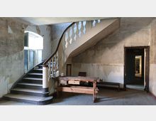 Ein Raum in einem alten Gebäude mit sichtbaren Gebrauchsspuren an den Wänden, einer geschwungenen Holztreppe mit weiß lackierten Balustern und einem großen Holzarbeitstisch im Vordergrund. Rechts führt eine Türöffnung in einen weiteren Raum.