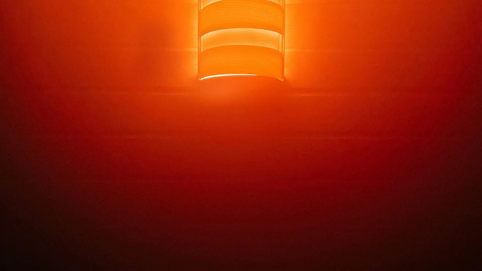 Innenansicht einer hölzernen Decke mit zentralem Deckenbalken, an dem eine zylindrische Deckenleuchte mit mehreren Lamellen hängt. Die Leuchte strahlt ein warmes orangefarbenes Licht aus und beleuchtet den Deckenbereich.