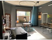 Ein kleiner Raum mit moderner Einrichtung und grauen Wänden, ausgestattet mit einem runden Tisch, schwarzen Stühlen und einem rechteckigen Tisch im Vordergrund. Große Fenster lassen Tageslicht einfallen, und links steht ein Regal mit Büchern und Postkarten; rechts im Bild befindet sich ein Modell auf einem Tisch.