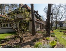 Ein zweigeschossiges Wohnhaus mit Satteldach und mehreren Gauben, umgeben von einem gepflegten Garten mit blühenden Beeten, hohen Bäumen und Rasenflächen. Im Vordergrund stehen Bäume und Sträucher, die den Blick auf die Hausfassade teilweise verdecken.