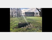 Eine ländliche Szene mit einem alten Backsteingebäude im Hintergrund, umgeben von Bäumen und einer grünen Wiese. Im Vordergrund befindet sich ein kleiner, aus Ziegelsteinen gebauter Wassergraben oder eine Struktur, die teilweise verfallen ist.