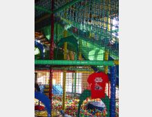 Indoor-Spielplatz mit Klettergerüsten, Netzen und einer bunten Bällebadfläche. Im Hintergrund sind grüne und gelbe Strukturen sowie große Fenster erkennbar, die den Bereich erhellen.
