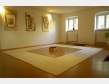 Ein minimalistischer, heller Raum mit hölzernem Boden, großen weißen Teppichen in rechteckiger Anordnung und einer kleinen Holzbox in der Mitte. An der Wand hängen abstrakte Kunstwerke, und die Umgebung wird durch zwei große Fenster mit Heizkörpern darunter belichtet.