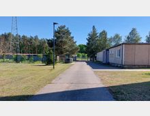 Gepflegter Hofbereich mit asphaltiertem Weg, flankiert von einem flachen Gebäude mit grauer Fassade auf der rechten Seite und einer Wiese auf der linken Seite. Im Hintergrund befinden sich ein Metalltor, ein Strommast und ein dichter Wald aus Nadelbäumen.