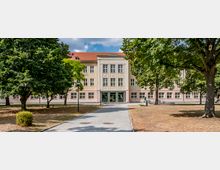 Ein dreigeschossiges Gebäude mit heller Fassade und rotem Ziegeldach, umgeben von Bäumen und einer gepflasterten Wegführung, die auf den Haupteingang zuführt. Der Vorderbereich umfasst eine parkähnliche Grünfläche mit Sitzbänken und einer Büste auf einem Sockel.