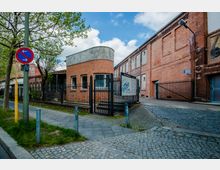 Backstein-Gebäude auf einem gepflasterten Hof mit einem Zaun und einem Eingangstor im Vordergrund. Links vorne ein Verkehrsschild, daneben Grünflächen und Bäume; im Hintergrund sind weitere Teile der Industriefassade sichtbar.