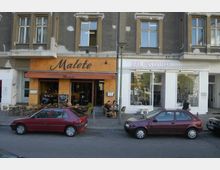 Straßenansicht eines Gebäudes mit einer orangefarbenen Café-Fassade, beschriftet "Malete", links, und einem weißen Laden mit großen Schaufenstern, beschriftet "GAL INSTITUTE", rechts. Vor dem Café stehen Tische und Stühle im Außenbereich, während einige parkende Autos die Straße säumen.