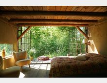 Schlafzimmer mit rustikaler Holzbalkendecke, großen Glaswänden und Blick auf üppigen, grünen Wald. Im Raum befinden sich ein Bett mit bunter Tagesdecke, zwei retro-inspirierte Sessel, eine Dekovase mit Blumen und Sitzkissen auf einem Holzfußboden, der in die Natur übergeht.