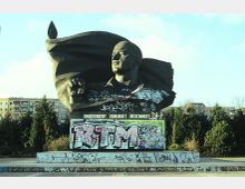 Denkmal einer männlichen Büste mit wehender Fahne, auf einem mit Graffiti bedeckten Sockel stehend; im Hintergrund sind moderne Wohngebäude und Bäume zu sehen. Text auf dem Sockel lautet: „Eingekerkert, Ermordet, Beschmiert“.
