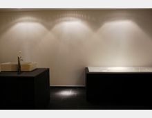 Moderner Badezimmerbereich mit dunklem Fliesenboden, einer schwarzen freistehenden Waschtischkonsole mit quadratischem Waschbecken und einer weißen Badewanne rechts. Die Wand ist schlicht und beige, beleuchtet durch mehrere Wandstrahler.