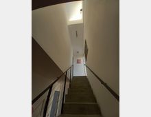 Betontreppe in einem modernen Innenbereich mit schlichtem weißen Wänden, metallischen Handläufen und einigen aufgehängten Kunstwerken; oben befindet sich ein rechteckiges Oberlicht, das natürliches Licht hereinlässt.