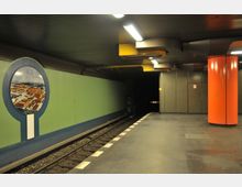 U-Bahnhof mit grün gestrichenen Wänden, einem rundem Bild in blauem Rahmen, einer orangefarbenen Säule und modern gestalteter Decke mit gelben Elementen. Links sind die Schienen und der Bahnsteig mit weißen Markierungen erkennbar.