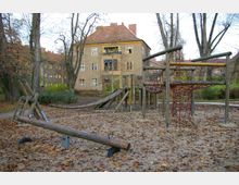 Spielplatz mit Holzklettergerüst, Wippe und Seilnetz auf einem mit Herbstlaub bedeckten Sandboden, umgeben von großen Bäumen. Im Hintergrund steht ein mehrstöckiges Wohnhaus mit gelben Fensterrahmen und einem roten Ziegeldach.