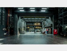 Blick auf den technischen Backstage-Bereich einer Theaterbühne mit sichtbaren Lagerräumen, Regalen mit Requisiten und Scheinwerfern, sowie verschiedenen Arbeitsmaterialien wie Leitern und Rollwagen. Der Raum ist dunkel gestaltet und von schwarzen Wänden und Stahlkonstruktionen umgeben.