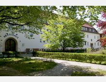 Ein weißes Ärztehaus mit roten Dachziegeln, großen Fenstern und einem Rundbogen-Eingang, umgeben von Bäumen, Hecken und einem gepflasterten Weg in einer grünen Parkanlage.