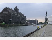Historische Hafengegend mit großen Backsteingebäuden, einem Kran und einer Wasserstraße, auf der kleinere Schiffe fahren; im Vordergrund eine gepflasterte Kaimauer mit Gleisen.