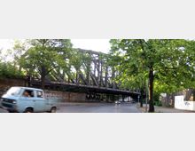 Eine alte Eisenbahnbrücke aus Metall überspannt eine gepflasterte Straße, flankiert von Bäumen und Graffiti-bedeckten Mauern. Im Vordergrund ist ein blauer Kleintransporter auf der Straße zu sehen.