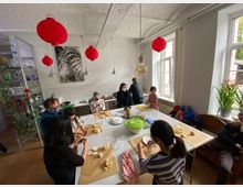 Eine Gemeinschaftsküche mit mehreren Personen, die an einem großen Tisch Plätzchen backen. Im Raum befinden sich helle Wände, rote Lampions an der Decke, große Fenster mit Pflanzen auf der Fensterbank und Regale mit Geschirr im Hintergrund.