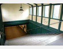 Treppenabgang in einem U-Bahn-Bereich mit grünen Metallträgern, cremefarbenen Wänden und kleinen Fenstern an den Seiten. Eine große Hängelampe beleuchtet den Bereich, und an den Wänden sind Graffiti sowie Plakate zu sehen.
