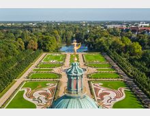 Blick auf einen barocken Schlossgarten mit symmetrisch angelegten Beeten, Rasenflächen und Ornamenten, eingerahmt von Baumreihen. Im Vordergrund steht die grüne Kuppel mit einer goldenen Statue auf einer Kugel, dahinter ein Fluss und bewaldete Umgebung.