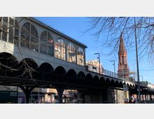 Überdachte Hochbahnbrücke mit großen Glasfenstern in bogenförmigen Rahmen, darunter Geschäftsfassaden und Straßenszene. Im Hintergrund ist ein roter Kirchturm mit spitzem Dach zu sehen.