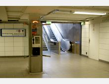 Unterirdischer Bahnhof mit einer Rolltreppe und Treppen, die nach oben führen. Links ist ein öffentlicher Münztelefonautomat an einer Säule angebracht, daneben ein Hinweisschild zu Bushaltestellen, und weiße Fliesenwände mit Türen prägen den Raum.