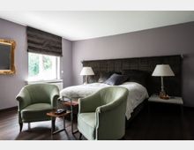 Schlafzimmer mit grauer Wandfarbe, einem großen Bett mit gepolstertem Kopfteil und weißen Bettlaken. Im Vordergrund stehen zwei grüne Sessel um kleine Beistelltische, während auf beiden Bettseiten Nachttische mit Tischlampen stehen; ein Fenster mit dunklem Rollo lässt Tageslicht herein.