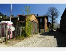 Pflastersteinstraße in einem Wohngebiet mit Backsteingebäuden, teils verfallen und mit Holztoren, daneben ein Zaun mit einer rosa mobilen Toilette. Im Hintergrund stehen große, unbelaubte Bäume und parkende Autos.