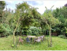 Ein Garten mit einer überdachten Pergola aus Metall und rankenden Pflanzen. Darunter steht eine Sitzgruppe aus mehreren Stühlen und einem Tisch; im Hintergrund sind weitere Pflanzen und Bäume zu sehen.
