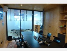 Ein kleiner Konferenzraum mit Holzwänden und einem dunklen Besprechungstisch mit mehreren Stühlen. Eine Glaswand mit halbtransparenter Sichtschutzfolie trennt den Raum von einem angrenzenden Bereich mit weißen Fliesenwänden und Schreibtischen.