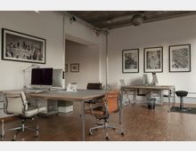 Moderner Büroraum mit hellen Wänden, Deckenbeleuchtung und Kunstwerken an den Wänden. Der Raum ist mit Schreibtischen, Bürostühlen und Computern ausgestattet, während der Holzboden eine warme Atmosphäre schafft.