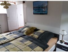 Ein Schlafzimmer mit einem Doppelbett, dessen Bettwäsche in Gelb- und Grautönen gemustert ist. Im Raum befinden sich zwei Nachttische mit Lampen, eine weiße Tür, ein kleines Fenster und ein Wandbild mit dem Motiv eines Yachtschiffs.