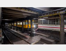 U-Bahn-Station mit gelbem Zug, der auf Schienen durch einen teilweise beleuchteten Tunnel fährt. Rechts sind das Stationsgebäude mit weißen Kacheln und einem roten Streifen sowie große Fenster zu sehen.