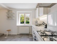 Helle, moderne Küche mit weißen Schränken, langer Arbeitsplatte und Gasherd unter einer Dunstabzugshaube. Ein großes Fenster über dem Heizkörper bietet Blick ins Grüne; links sind offene Wandregale und ein kleiner Hocker.