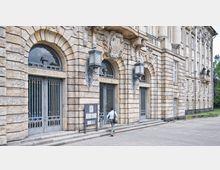 Historische Steinfassade eines Gebäudes mit großen, verzierten Rundbögen, Metallgittertüren und dekorativen Elementen oberhalb der Eingänge. Vor dem Eingang geht eine Person, und links steht ein Schild mit Informationen.