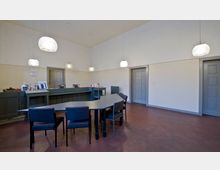 Ein schlicht gestalteter Büro- oder Empfangsraum mit einem grauen Tresen, einem schwarzen Tisch mit mehreren Stühlen und einer weißen Wand. Im Raum hängen mehrere kugelförmige Deckenleuchten, und graue Türen sind gut sichtbar in den Wänden integriert.