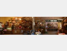 Ein historisch eingerichteter Tante-Emma-Laden mit hölzernen Regalen, die mit alten Verpackungen, Gläsern und Dosen gefüllt sind. Im Vordergrund stehen eine Glasvitrine mit Süßigkeiten, Kisten mit Waren und ein Verkaufsbereich mit antiker Kasse.