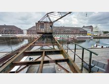 Historischer Hafenbereich mit einem alten, verrosteten Kran im Vordergrund, der auf Schienen montiert ist. Im Hintergrund sind Lagerhäuser aus Backstein, geparkte Schiffe und ein Kanal zu sehen.