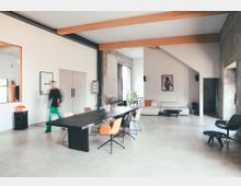 Geräumiger Wohn- und Essbereich mit modernen, minimalistischen Möbeln, darunter ein langer Holztisch mit bunten Stühlen und eine helle Eckcouch. Sichtbare Holzbalken, eine teils rohe Betonwand, große Fenster und dezente Dekorelemente prägen den Raum.