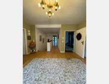 Flur in einem Wohnhaus mit Holzboden und hellem Teppich. Im Vordergrund hängt ein goldfarbener Kronleuchter, während im Hintergrund ein Flur mit weißen Wänden, Kunstwerken und Fenstertüren sichtbar ist. Links und rechts befinden sich Türen zu weiteren Räumen.