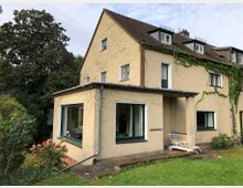 Zweistöckiges Wohnhaus mit beigem Putz, begrüntem Satteldach und teilweise bewachsener Fassade. Im Vordergrund befinden sich ein Garten mit Rasen, Blumen und Sträuchern sowie eine Veranda mit einem Liegestuhl.