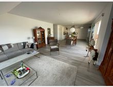 Wohnzimmer mit offener Küche in modernem Stil. Zu sehen sind ein graues Sofa, ein Teppich, ein Couchtisch mit Dekoration, eine Essgruppe mit Holzstühlen, sowie ein dekoratives Schaukelpferd und ein antiker Holzschrank; der Raum hat helle Wände und Laminatboden.