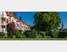 Reihen von roten Backstein-Wohnhäusern mit weißen Balkonen, umgeben von gepflegten Gärten und Büschen; im Hintergrund ragt ein Kirchturm mit grünem Spitzdach hervor. Ein großer Baum und ein Maschendrahtzaun trennen die Gebäude von einer Grünfläche.