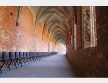 Ein gotischer Kreuzgang mit roten Backsteinwänden, spitzbogigen Gewölben und seitlich angeordneten schwarzen Plastikstühlen; der Boden ist mit grauen Fliesen ausgelegt.