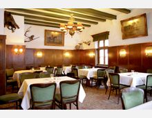 Innenraum eines traditionellen Restaurants mit Holzwänden, grünen Polsterstühlen und weiß eingedeckten Tischen. An den Wänden hängen Gemälde, Jagdtrophäen und dekorative Beleuchtungen, während eine große Fensterfront Tageslicht hereinlässt.