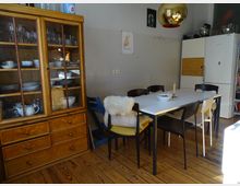 Esszimmer mit Holzboden, einem weißen rechteckigen Tisch und schwarzen Stühlen, teilweise mit Schaffellauflagen. Links steht ein rustikaler Holzschrank mit Glasvitrine, rechts ein Kühlschrank und Regal mit Küchengeräten, darüber hängt eine goldene Lampe.