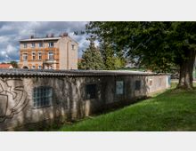 Ein Flachbau mit Wellblechdach und abgenutzter Außenwand, die mit vereinzeltem Graffiti bedeckt ist, steht vor einem dreigeschossigen Wohngebäude aus Ziegeln und Putz. Im Vordergrund ist eine Rasenfläche mit einem großen Baum, im Hintergrund sind weitere Bäume und Gebäude sichtbar.